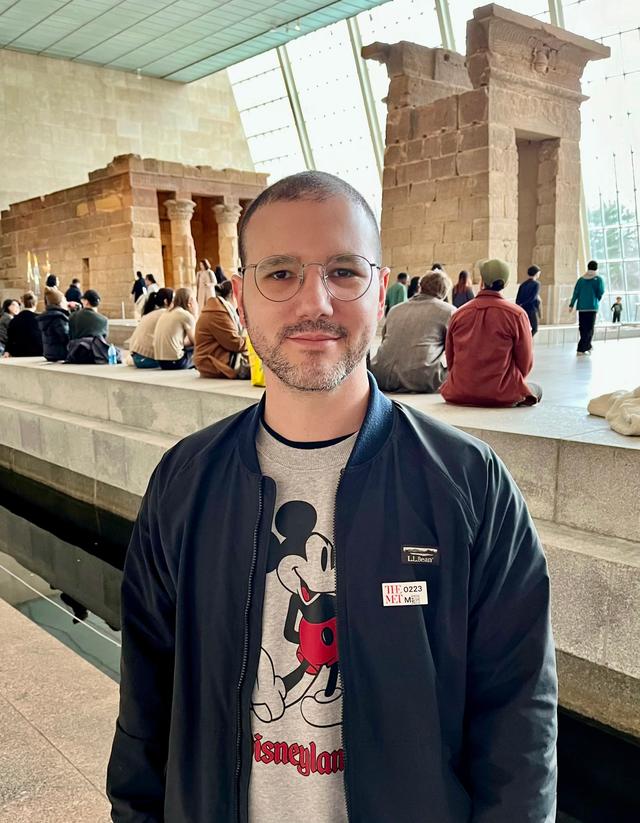 A man in a gray Mickey Mouse sweater stands in front of the Temple of Dendur at the Metropolitan Museum of Art in New York City.