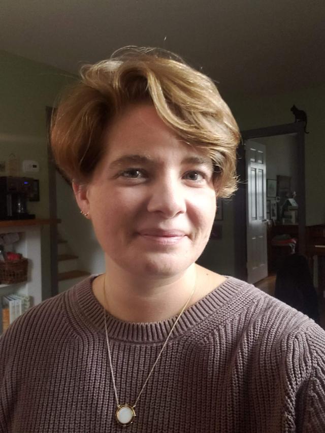 Photo from the shoulders up of a white woman in her 30s with short blonde hair. She is smiling with a closed mouth and wearing a light brown sweater.