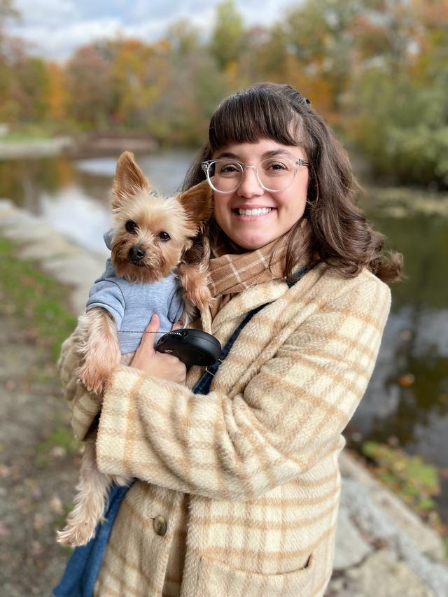Photo of a white woman with shoulder length brown hair, wearing clear frame glasses, a tan and beige plaid coat, and holding a small brown dog in grey sweatshirt. 